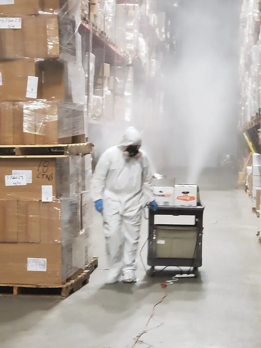 A Marrone technician in a protective suit fogging a warehouse aisle beside racks of stacked pallets